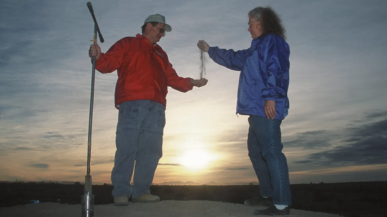 Two people taking soil samples with a probe at sunset
